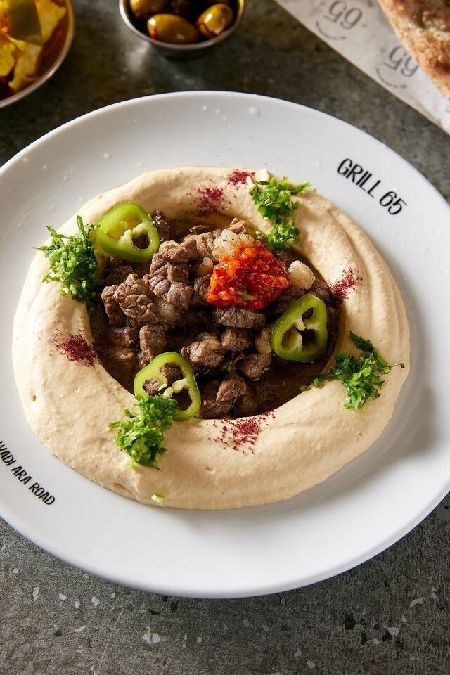 צלחת חומוס עם ערימת קוביות בשר צלוי במרכז, פלפל ירוק חריף וסלסה אדומה בצד